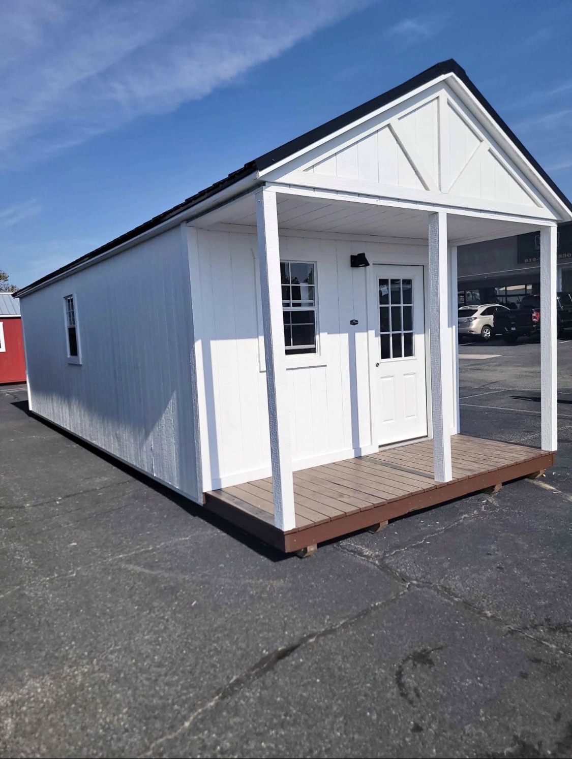 White portable cabin with a black gabled roof, wooden front porch with white posts, and a windowed door on an asphalt lot
