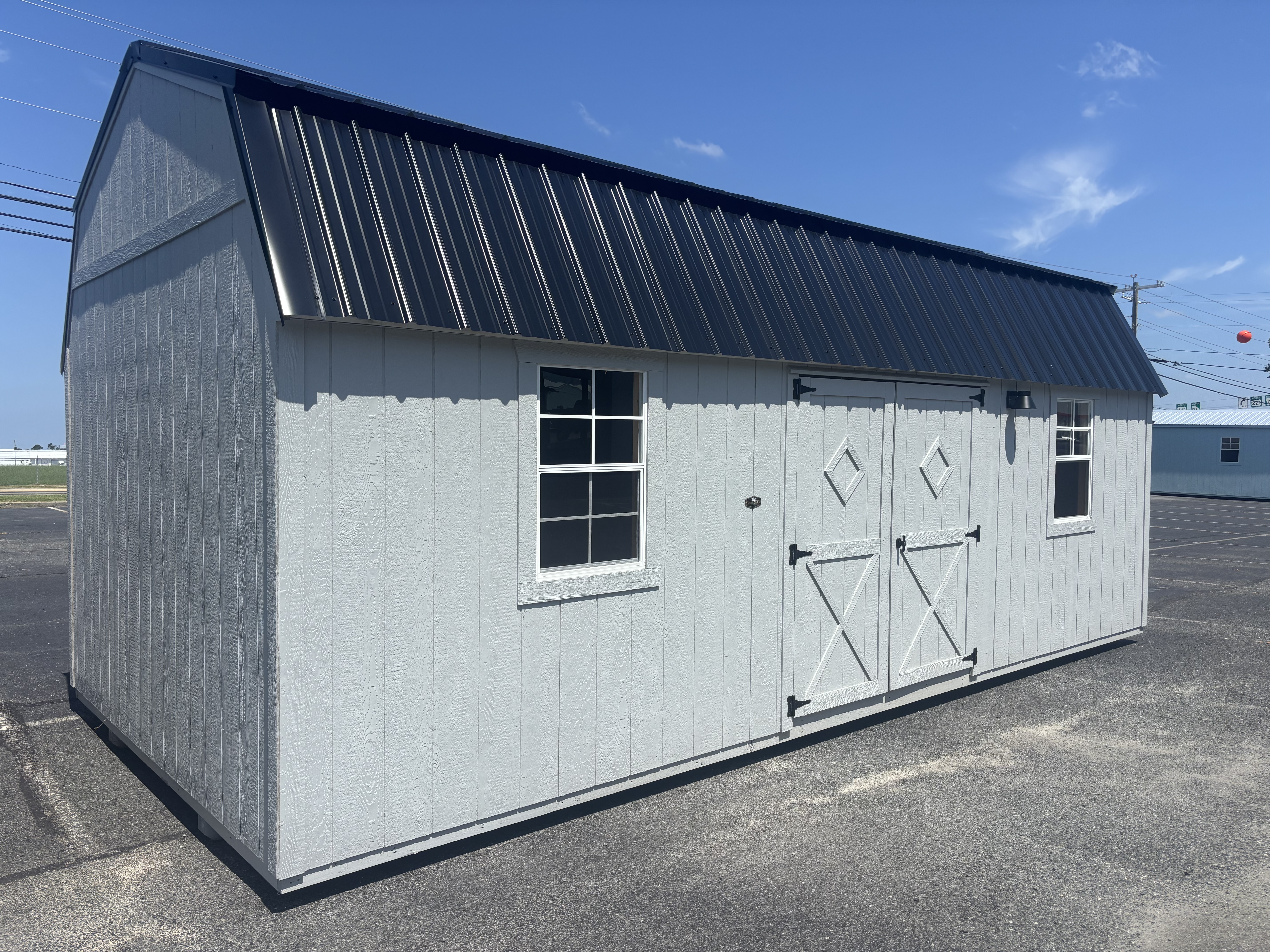 Light gray lofted barn portable building with barn-style double doors and a black roof on a sandy lot