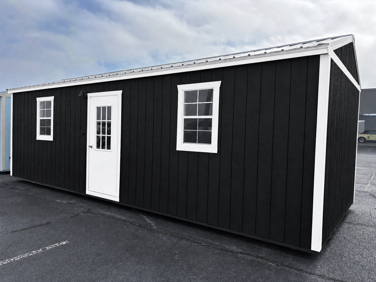 Black utility shed with vertical board-and-batten siding, white trim, a single white door, and a low-slope gable roof on an asphalt lot